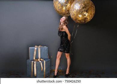 Enthusiastic white girl with sparkle helium balloons enjoying  birthday photoshoot on dark background. Adorable female model in black dress posing in studio with big present boxes and smiling. - Powered by Shutterstock