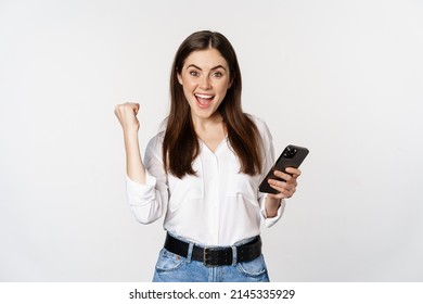 Enthusiastic woman winning on mobile phone, rejoicing and screaming of joy while using smartphone app, standing over white background - Shutterstock ID 2145335929