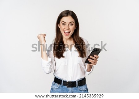 Enthusiastic woman winning on mobile phone, rejoicing and screaming of joy while using smartphone app, standing over white background