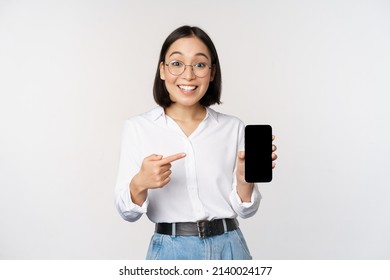 Enthusiastic young asian woman pointing finger at smartphone screen, showing advertisement on mobile phone, white background - Shutterstock ID 2140024177