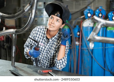 Enthusiastic young female welder in casual plaid shirt, protective gloves and helmet welding metal pipe structure in workshop, holding torch and carefully inspecting quality of weld seams - Powered by Shutterstock