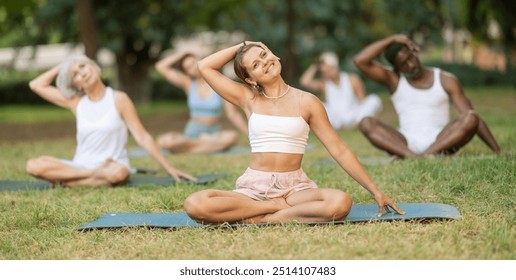 Enthusiastic young female yoga instructor leading outdoor group class in tranquil setting amidst lush greenery of city park, performing stretching exercises in Padmasana pose.. - Powered by Shutterstock