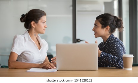 Enthusiastic young indian employee sharing new project ideas with serious female boss. Diverse business woman team discussing working moments at office. HR manager listening to interns experience