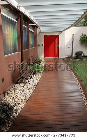 Entrada externa com deck de madeira, bancos laterais e porta vermelha, em espaço moderno com jardim e cobertura translúcida.