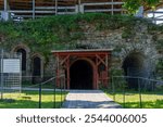 Entrance to an ancient stone fortress with a wooden walkway surrounded by green trees. The historic architecture evokes a sense of medieval heritage and tranquility