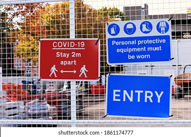 Entrance To A Construction Site With Warning Signs Attached To A Fence. Covid 19 Stay 2 Metres Apart. Personal Protective Equipment Must Be Worn. Entry.