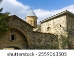 Entrance to the courtyard of the Kvatakhevi monastery. Stone walls, metal gates, arch, tower, dome of the church. Bright blue sky with clouds.