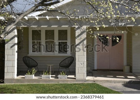 ENTRANCE WITH COVERED PORCH VERANDAH AND PRETTY PINK FRONT WOODEN DOORS  - An old beige period character style timber siding board home with leadlight windows, a patio awning, table, chairs and garden