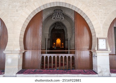 The entrance to the Great Mosque of Sousse, Tunisia, features a grand wooden door and intricate archways. The warm, golden light from inside creates a sense of mystery and invites visitors to explore. - Powered by Shutterstock