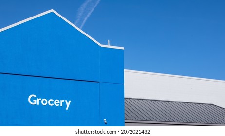 Entrance To A Grocery Store. White Text On Blue Wall With Pointy Triangular Roof. Grainy Stucco Architecture, A Popular Texture Found In Southern Nevada. Clear Sky With A Single Airplane Cloud Above.