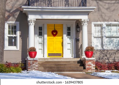 Entrance Of Pretty Vintage House With Ornate Columns On Porch And Red Valentine Wreath On Bright Yellow Door In Snow