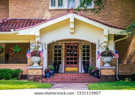 Entrance and stuco bungalow porch with french doors and tile roof on brick house with many flower pots and American flags and star shaped door wreath
