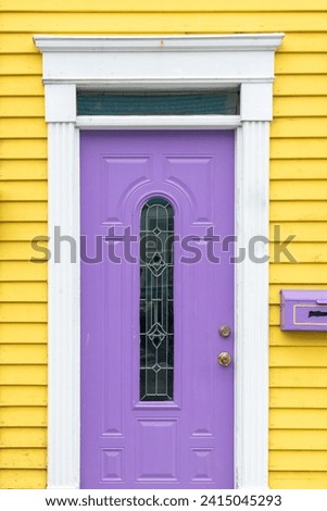The entrance to a vibrant yellow wooden house with clapboard siding and a purple mailbox. There's a single purple metal door with a narrow glass decorative window, white trim and a transom window.