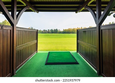 Entrance with wooden fence and roof to modern golf driving range with big green field and trees under cloudy sky on sunny day - Shutterstock ID 2251937979