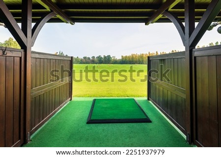 Entrance with wooden fence and roof to modern golf driving range with big green field and trees under cloudy sky on sunny day
