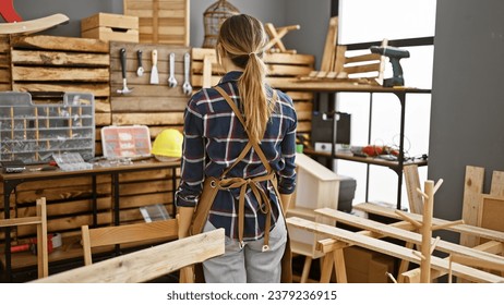 Entrancing back view of a young blonde carpenter, standing backwards, wearing her apron amidst a buzzing carpentry workshop - Powered by Shutterstock