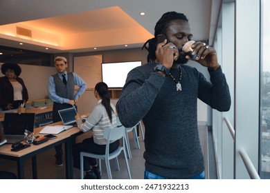 An Entrepreneur black Man with dreadlocks is drinking coffee an listening to the phone call standing by the window in the office. Is wearing a sweater and a watch, there are other people in the room - Powered by Shutterstock