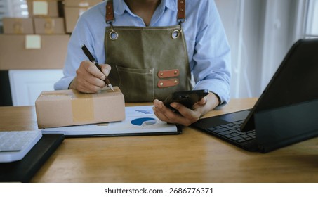 Entrepreneur checking smartphone while preparing a package for shipping, working with laptop, documents, and calculator on desk.
