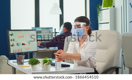 Entrepreneur with protection mask and visor applying sanitizer gel rubbing hands before typing at computer. Businesswoman in new normal workplace disinfecting while colleagues working in background
