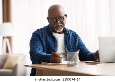 Entrepreneurship Career. Mature African American Man Taking Notes Working Sitting At Laptop Computer In Modern Office Indoors. Senior Businessman Writing Report At Workplace, Wearing Eyeglasses
