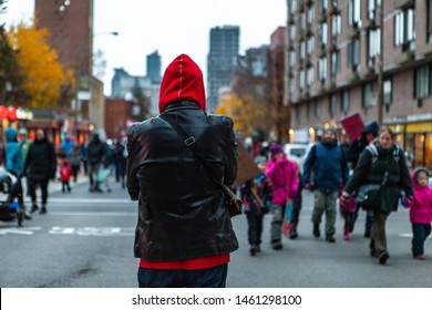 Environmental activists march in city. A bystander watches on as a large crowd of ecological protestors marches towards the camera, person wearing a hood from behind with blurred street in background