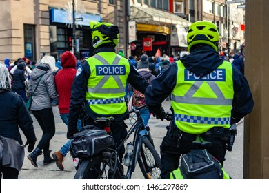 Environmental Activists March In City. Two On Duty Police Officers Wearing High Visibility Uniforms Are Seen From Behind With Pedal Bikes, Watching People For Public Safety During A Street Rally.