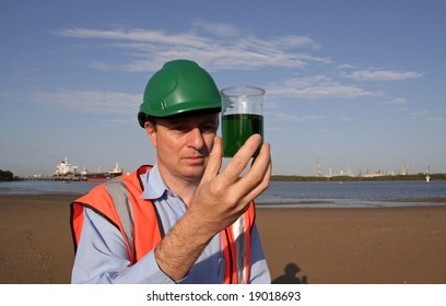 An Environmental Engineer On The Mudflats Examining A Sample Of Oil From The Ship Docked Behind Him, Showing The Estuary And Beautiful Blue Sky, Wearing Orange Reflective Vest And Green Safety Helmet