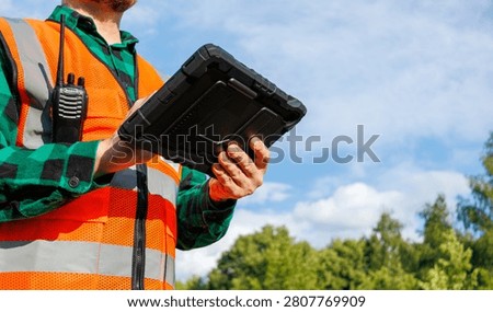 Environmental engineer or site inspector holding a rugged digital tablet and wearing an orange reflective safety vest with a walkie-talkie. Technician or ecologist using a smart device for data collection and fieldwork operations against a background of green trees and blue sky.