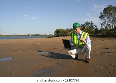 An environmental engineer, wearing protective clothing on his laptop computer analyzing data from the  mudflats in the estuary.