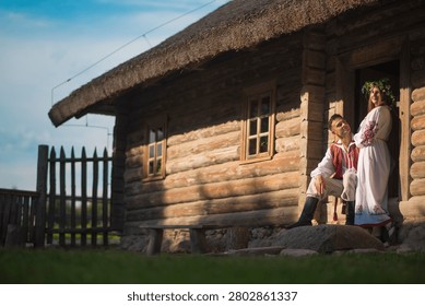 Environmental horizontal portrait of a young eastern european man and woman in traditional vyshyvanka dresses posing near a historic log cabin in warm sunset light