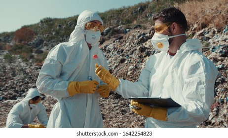 Environmental Pollution Issue. Team Of Professional Ecologists Researching Dump In Sunny Weather. Two Scientists Examine Toxic Sample And Write Down Results.