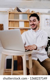 Environmental Portrait Of A Happy Smiling Handsome Middle Aged Brunet Furniture Designer Maker In His Workshop Behind Handmade Stool With A Laptop. Wood Samples And Pictures Hanging On The Wall.