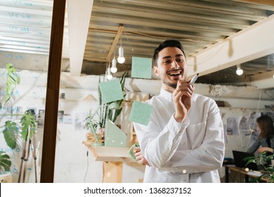 Environmental Portrait Of A Happy Smiling Handsome Middle Aged Brunet Furniture Designer Maker In His Workshop Through Window. He Is Drawing Important Measurements And Markings On His Glass Board.