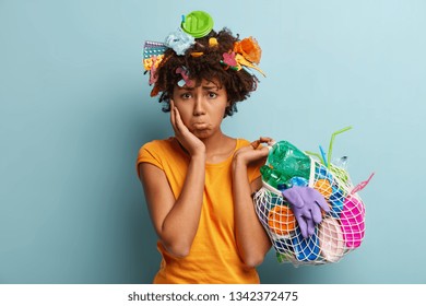 Environmental problems concept. Dissatisfied dark skinned woman with displeased dejected expresses, tries to remove plastic from Earth, takes products for recycling, isolated on blue wall. Earth Day