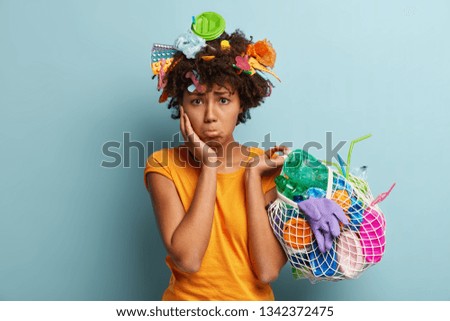 Environmental problems concept. Dissatisfied dark skinned woman with displeased dejected expresses, tries to remove plastic from Earth, takes products for recycling, isolated on blue wall. Earth Day