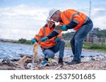 Environmental scientists or workers, wearing safety helmets and gloves, examining waste materials along a shoreline are large industrial tanks environmental waste water or pollution effects concept.