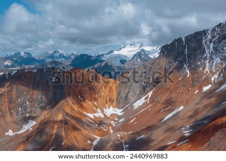Epic aerial view to big sharp rocky ridge of red color and huge snow-capped mountain peak hidden in low cloudiness. Beautiful giant snow mountain top in cloudy sky. Shadows of clouds on colorful rocks