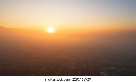 Epic gray smog is visible at sunset over the city. A bird's-eye view from a drone of houses, roads, cars and parks. White clouds and snowy mountains are illuminated by orange rays of the sun. Almaty - Powered by Shutterstock