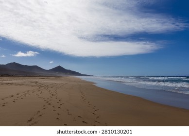 An epic landscape capturing a wide, empty shoreline with human footprints leading towards a dramatic mountain range. The composition beautifully balances the power of the ocean, the grand scale of the mountains, and the feeling of personal freedom and exploration.