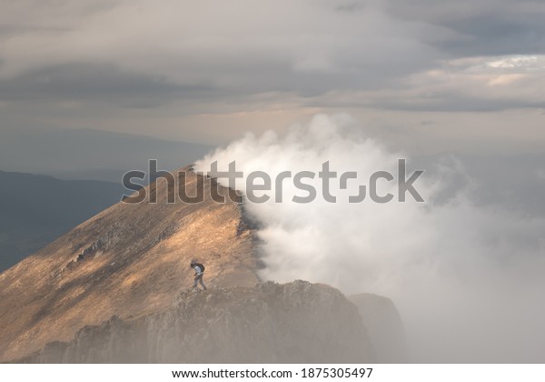 Epic view of mountain hiker\
standing on the top of the cliff and distant mountain ridge lighten\
by soft sunlight from one side and covered by thick fog from\
another