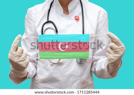 Epidemic in Azerbaijan. Young woman doctor in a medical coat (suit) and gloves holds a medical mask with the print of the flag of Azerbaijan on a blue background isolated.