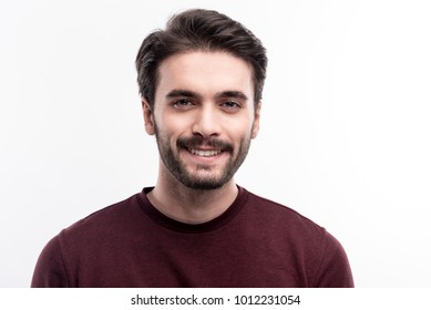 Epitome of masculinity. The portrait of a handsome dark-haired bristled young man in a burgundy sweater smiling at the camera while posing against a white background