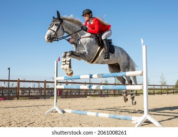 An equestrian athlete jumps a high barrier on a horse. The girl is dressed in a red competition jacket and white breeches.On the athlete's head is a protective helmet. Image with selective focus - Powered by Shutterstock