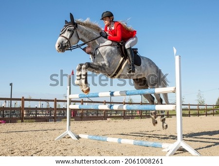 An equestrian athlete jumps a high barrier on a horse. The girl is dressed in a red competition jacket and white breeches.On the athlete's head is a protective helmet. Image with selective focus
