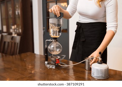 Equipment, Coffee Shop, People And Technology Concept - Close Up Of Woman With Butane Gas Burner Heating Water In Siphon Coffeemaker At Cafe Bar Or Restaurant Kitchen