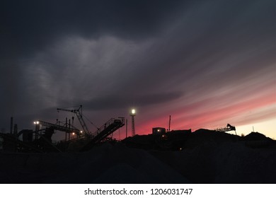 Equipment for crushing rock and sorting into fractions at a mining enterprise. Silhouettes of constructions against the background of red evening sky.
