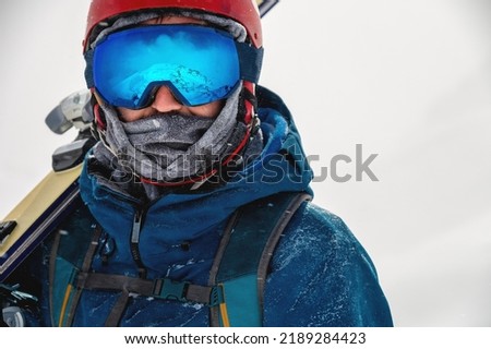 Equipped skier holding skis on his shoulder and looking directly at the camera, portrait. Man with winter equipment in the mountains at a ski resort