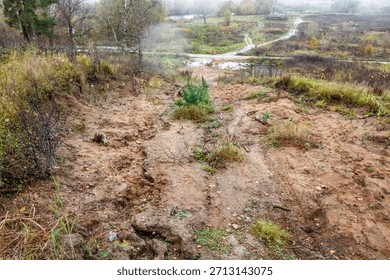Eroded hillside with sandy soil and sparse autumn vegetation overlooking a misty landscape with wet fields and distant trees