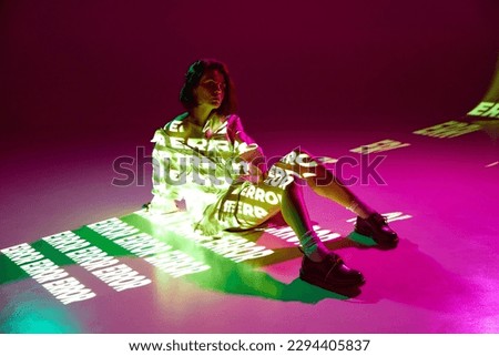 Error, mistake. Portrait of dreaming girl sitting on floor with digital neon filter lights with inscryption on body on pink mode background. Concept of digital art, fashion, cyberpunk, futurism