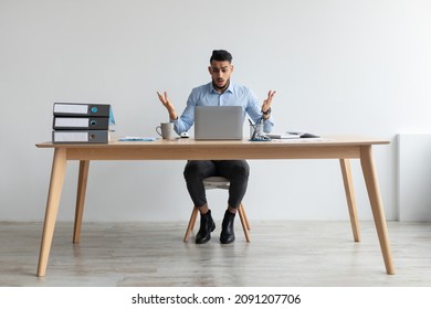 Error. Portrait of confused Arab guy sitting at desk with laptop, shrugging shoulders spreading hands. Remote Work. Stressed man suffering business problems, reading unexpected bad negative news